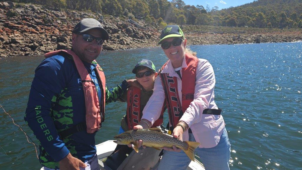 Three anglers in a boat on a lake, one holding a wild trout